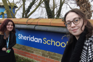 two trainee teachers standing beside a sign outside of a school