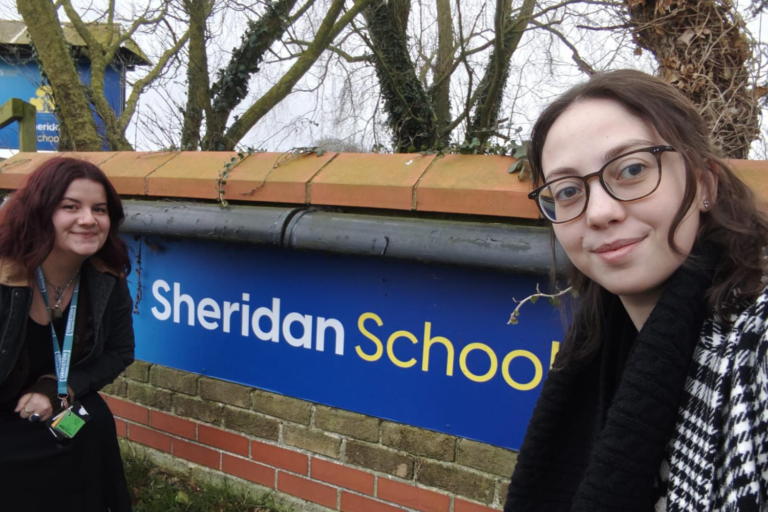 two trainee teachers standing beside a sign outside of a school